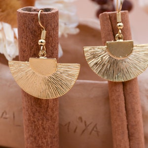 May include: A pair of gold-toned earrings with a fan-shaped design. The earrings are hanging from a cinnamon stick.