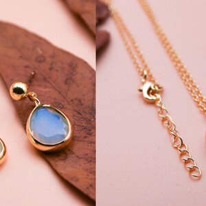 May include: A gold necklace and earrings set with a teardrop-shaped opal pendant. The opal is a light blue color with a white iridescent sheen.