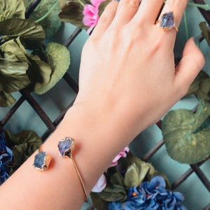 Puede incluir: Una pulsera de oro con dos acentos de cristal morado oscuro. La pulsera est&aacute; en la mu&ntilde;eca de una persona. La persona lleva un anillo de oro con un acento de cristal morado oscuro en su dedo.
