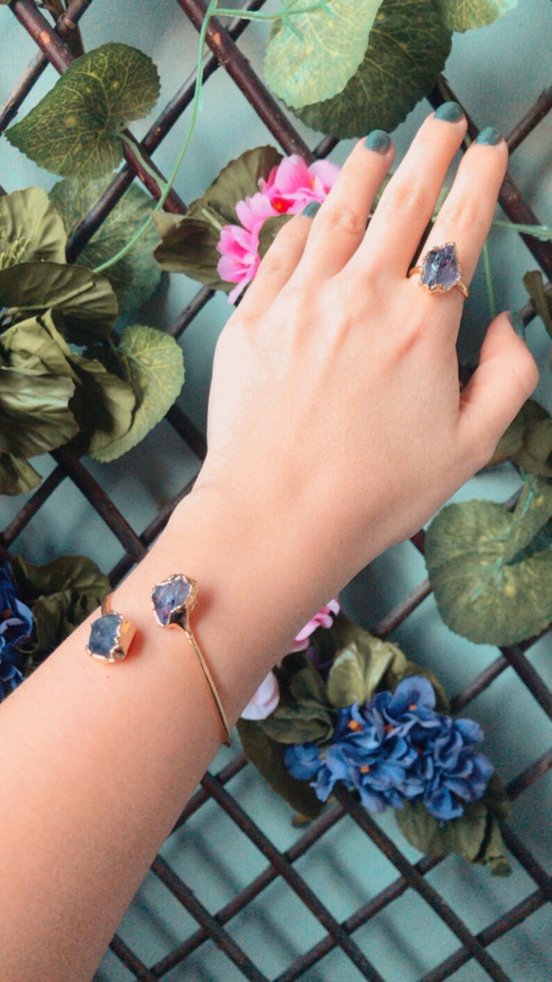Puede incluir: Una pulsera de oro con dos acentos de cristal morado oscuro. La pulsera est&aacute; en la mu&ntilde;eca de una persona. La persona lleva un anillo de oro con un acento de cristal morado oscuro en su dedo.