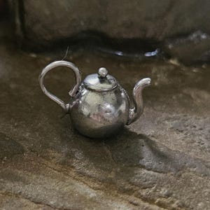 May include: A miniature silver-toned metal teapot charm featuring a rounded body, a curved handle, a small spout, and a lid with a tiny spherical knob on top. The surface of the teapot has a polished metallic finish with subtle reflective highlights.