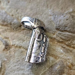 Peut inclure: Pendentif argenté détaillé avec une surface texturée. Le pendentif présente un design unique, peut-être une figure ou un objet stylisé. Il est posé sur un fond neutre en forme de pierre, soulignant ses détails complexes.
