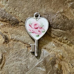 May include: A silver-toned metal charm shaped like a key with a heart-shaped bow. The heart features a white textured enamel inlay decorated with a delicate pink floral pattern and small green leaves. A small circular loop is at the top.