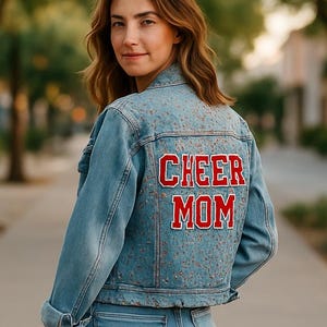 May include: Light blue denim jacket with the words "CHEER MOM" in red lettering on the back. The jacket is worn with matching blue jeans. The image is taken outdoors with trees and a sidewalk in the background.