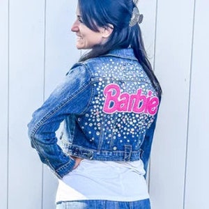 Può includere: Una giacca di jeans blu con un logo "Barbie" rosa decorato con strass sul retro.