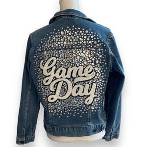 May include: A blue denim jacket with a white "Game Day" design on the back, embellished with rhinestones.