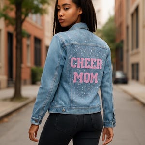 Pode incluir: Casaco de ganga azul claro com a inscrição rosa "CHEER MOM" nas costas. O casaco tem um design clássico com botões e bolsos. A parte de trás do casaco é decorada com glitter prateado.