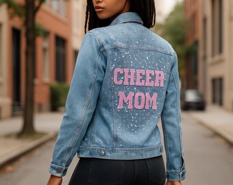 May include: Light blue denim jacket with the words "CHEER MOM" in pink lettering on the back. The jacket has a classic design with a button-down front and pockets. The back of the jacket is decorated with silver glitter.