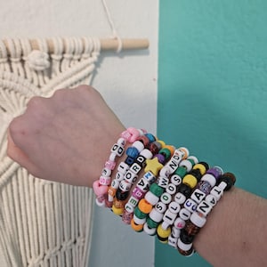 May include: A collection of colorful beaded bracelets spelling out names and phrases. The bracelets feature various colors and patterns, with white letter beads. The bracelets are stacked on a wrist.