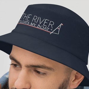 May include: A navy blue bucket hat with the text "THE RIVER FLOATS MY BOAT" in white and a red and white sailboat graphic.