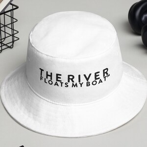以下が含まれることがあります： 黒い刺繍で「THE RIVER FLOATS MY BOAT」と書かれた白いバケットハット。