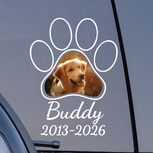 May include: A white paw print car decal with a photo of a golden retriever dog inside. The dog's name, "Buddy", and the years "2013-2026" are printed below the paw print in white script.