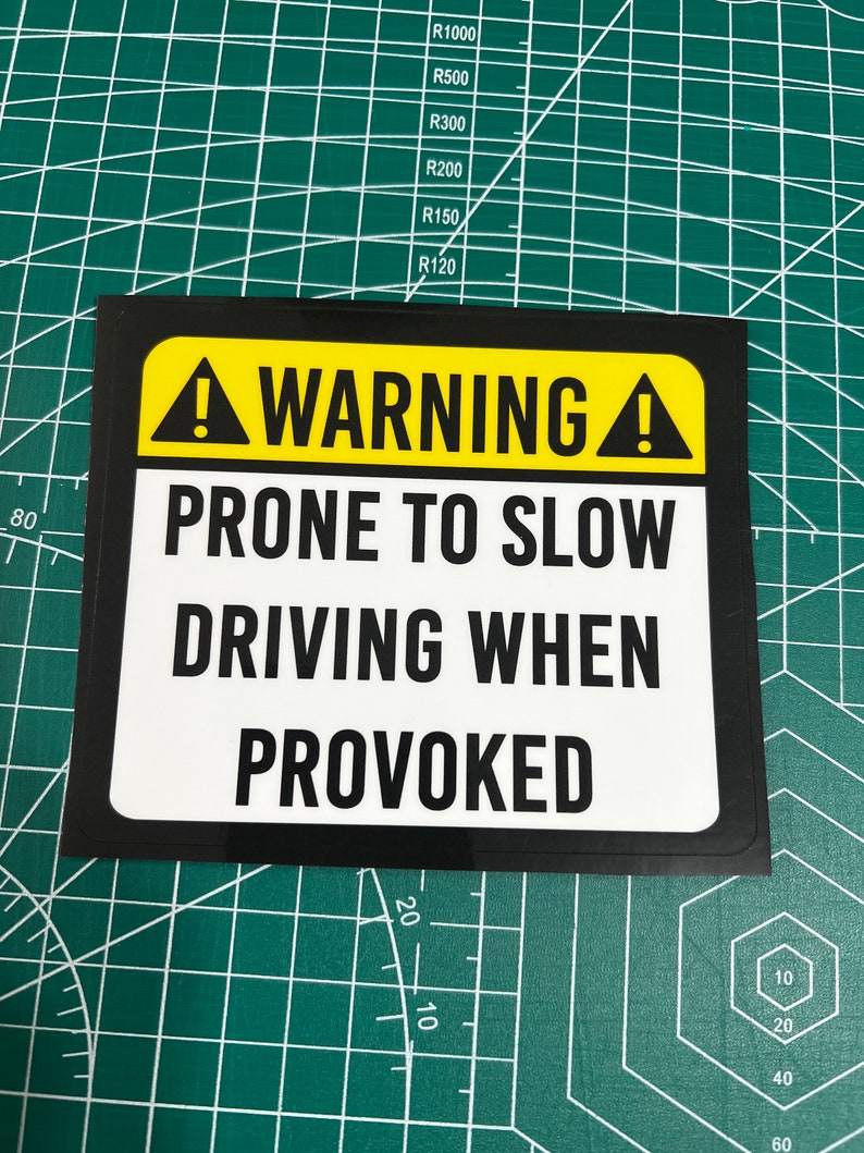 May include: A black and yellow warning sign with white text that reads "WARNING! PRONE TO SLOW DRIVING WHEN PROVOKED".