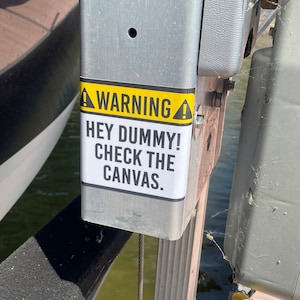 May include: A yellow and black warning sign with the text "WARNING! HEY DUMMY! CHECK THE CANVAS." attached to a metal post.