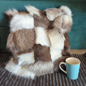 Könnte beinhalten: Ein quadratisches Zierkissen mit einem Patchwork-Design aus braunem, weißem und beigem Fell. Das Kissen liegt auf einer strukturierten, braunen Decke. Eine hellblaue Tasse mit weißem Henkel und goldenem Rand steht daneben.