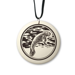 May include: A white ceramic pendant with a black and white illustration of a manatee swimming in the water. The pendant is on a black cord.