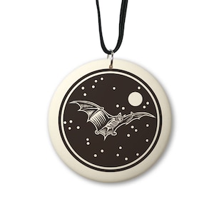 May include: A round, cream-colored ceramic pendant with a black cord. The pendant features a black and white illustration of a bat flying in front of a full moon and stars.