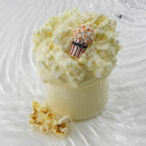Könnte beinhalten: Ein skurriles Dessert, das einem Popcorn-Snack ähnelt. Die cremige, cremeweiße Basis ist mit einer flauschigen, popcornartigen Substanz bedeckt, die mit einer Miniatur-Popcorntüte verziert ist. Ein paar Popcornstücke sind in der Nähe verstreut. Das Gesamtthema ist Essen und Süßigkeiten.