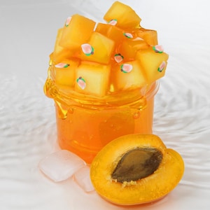 Könnte beinhalten: Ein klares Glas gefüllt mit orangefarbener Flüssigkeit und bedeckt mit gelben Würfeln, die mit kleinen Pfirsichmotiven verziert sind. Eine halbierte Aprikose mit sichtbarem Kern und zwei Eiswürfel befinden sich vor dem Glas, alles auf weißem Untergrund.