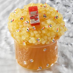 Puede incluir: Un recipiente de plástico transparente lleno de slime naranja, cubierto con cuentas amarillas translúcidas y pequeños dijes en forma de manzana. Un cartón rojo en miniatura con la etiqueta Apple Juice 70ml 100% descansa sobre las cuentas y el slime.