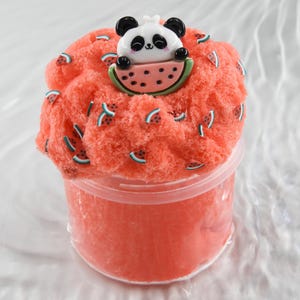 Puede incluir: Un recipiente de plástico transparente lleno de slime rojo con rodajas de sandía incrustadas. Una pequeña figura de panda está sentada encima del slime, sosteniendo una rodaja de sandía.