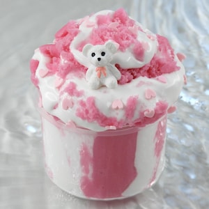 Könnte beinhalten: Ein durchsichtiger Kunststoffbehälter gefüllt mit einem rosa und weißen Dessert. Das Dessert hat eine weiße Schlagsahne mit rosa Streuseln und eine kleine weiße Teddybärfigur. Rosa Herzen verzieren den Rand.