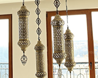 May include: An elaborate brass chandelier featuring four cylindrical lamps with a pierced pattern. The lamps are suspended from a geometric frame. The light fixture hangs from the ceiling, with a light-coloured wall and window in the background.