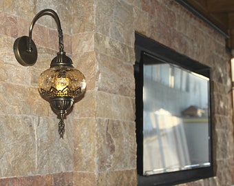 May include: An ornate, antique-style wall sconce with a curved arm and a decorative, globe-shaped glass shade. The shade has a textured, crackled appearance and intricate metalwork. The sconce is mounted on a stone-tiled wall.
