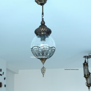 May include: A clear glass globe pendant light with intricate metalwork details hangs from a chain. The globe is frosted and has a decorative metal finial at the bottom. The light is suspended from a ceiling in a room with white walls.