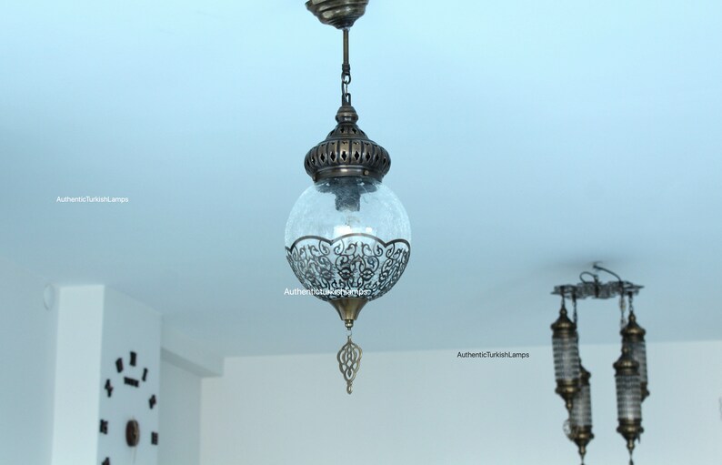 May include: A clear glass globe pendant light with intricate metalwork details hangs from a chain. The globe is frosted and has a decorative metal finial at the bottom. The light is suspended from a ceiling in a room with white walls.