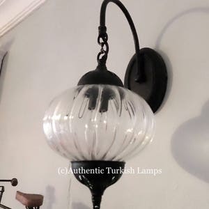 May include: A black metal wall sconce with a clear glass globe shade. The sconce has a curved arm and a round base. The glass shade has a ribbed texture. (c) Authentic Turkish Lamps