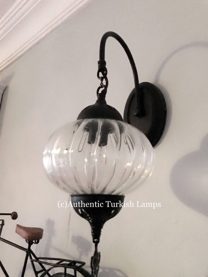 May include: A black metal wall sconce with a clear glass globe shade. The sconce has a curved arm and a round base. The glass shade has a ribbed texture. (c) Authentic Turkish Lamps