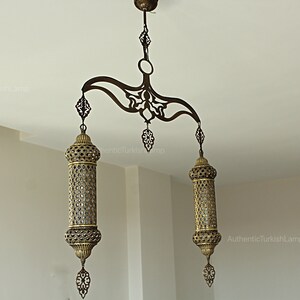 May include: An ornate bronze Turkish chandelier with two cylindrical lamps and decorative filigree. The lamps hang from a curved, patterned support attached to the ceiling. The text "AuthenticTurkishLamp" is visible.