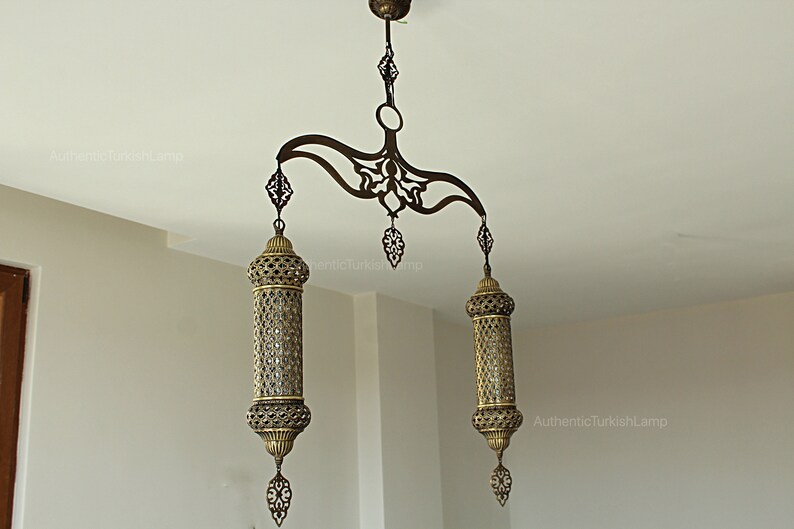 May include: An ornate bronze Turkish chandelier with two cylindrical lamps and decorative filigree. The lamps hang from a curved, patterned support attached to the ceiling. The text "AuthenticTurkishLamp" is visible.