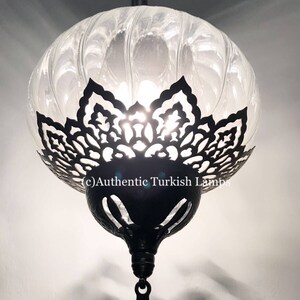 May include: A clear glass Turkish lamp with a black metal frame and intricate cutouts. The lamp is hanging from a chain and has a decorative finial at the bottom.