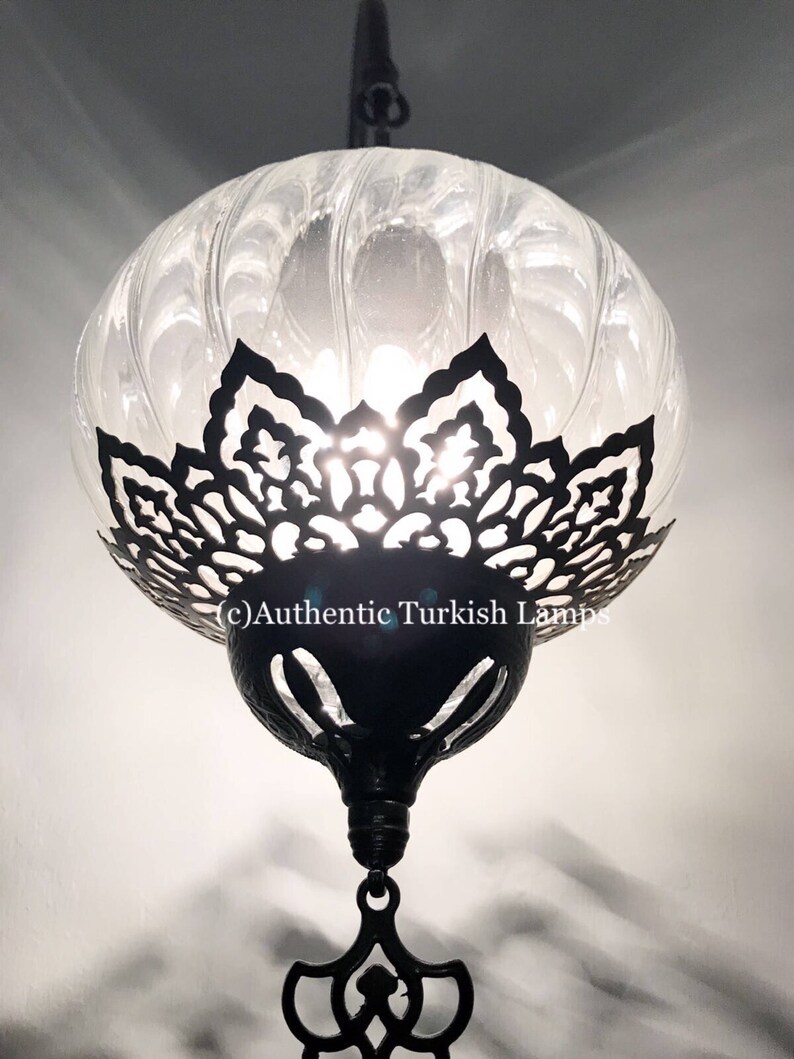 May include: A clear glass Turkish lamp with a black metal frame and intricate cutouts. The lamp is hanging from a chain and has a decorative finial at the bottom.