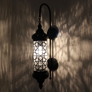 May include: A black metal wall sconce with a clear glass shade featuring a geometric pattern. The light is on and casting a warm glow.