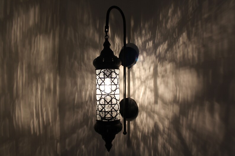 May include: A black metal wall sconce with a clear glass shade featuring a geometric pattern. The light is on and casting a warm glow.