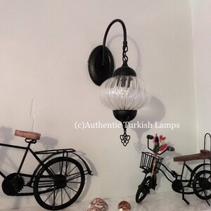 May include: A black metal wall sconce with a clear glass globe shade. The sconce is mounted on a white wall and has a decorative metal design at the bottom. The image also shows a bicycle and seashells on a shelf below the sconce. (c) Authentic Turkish Lamps