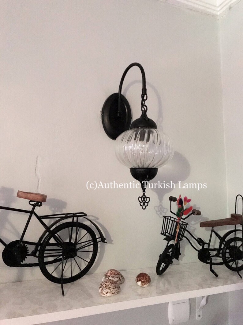 May include: A black metal wall sconce with a clear glass globe shade. The sconce is mounted on a white wall and has a decorative metal design at the bottom. The image also shows a bicycle and seashells on a shelf below the sconce. (c) Authentic Turkish Lamps
