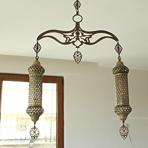 May include: An ornate, antique-style hanging lamp with a bronze finish. The lamp features a decorative, curved top with intricate cut-out designs, supporting two cylindrical, open-work shades. Each shade has a decorative finial.