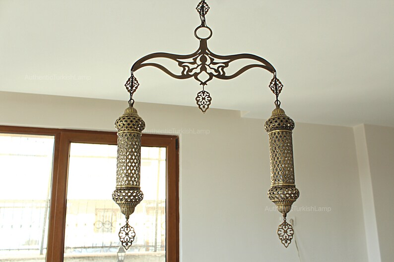 May include: An ornate, antique-style hanging lamp with a bronze finish. The lamp features a decorative, curved top with intricate cut-out designs, supporting two cylindrical, open-work shades. Each shade has a decorative finial.