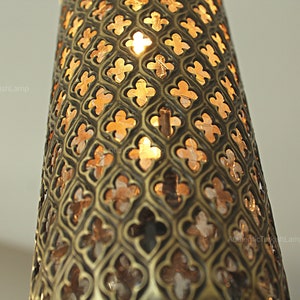 Puede incluir: Primer plano de una l&aacute;mpara cil&iacute;ndrica decorativa con un patr&oacute;n calado detallado. El metal de la l&aacute;mpara tiene un acabado bronce y el dise&ntilde;o recortado permite que la luz c&aacute;lida brille. La est&eacute;tica general recuerda al dise&ntilde;o turco o de Oriente Medio.