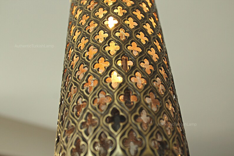 Puede incluir: Primer plano de una l&aacute;mpara cil&iacute;ndrica decorativa con un patr&oacute;n calado detallado. El metal de la l&aacute;mpara tiene un acabado bronce y el dise&ntilde;o recortado permite que la luz c&aacute;lida brille. La est&eacute;tica general recuerda al dise&ntilde;o turco o de Oriente Medio.