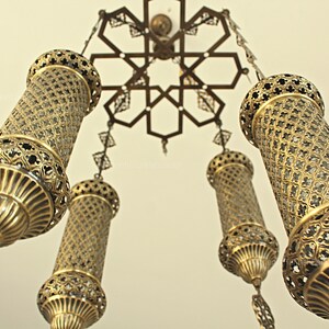 Puede incluir: L&aacute;mpara de ara&ntilde;a antigua y ornamentada con cinco l&aacute;mparas cil&iacute;ndricas doradas. Las l&aacute;mparas presentan intrincados dise&ntilde;os de metal calado y cuelgan de un accesorio de techo decorativo con un patr&oacute;n en forma de estrella. La est&eacute;tica general es vintage y elegante.