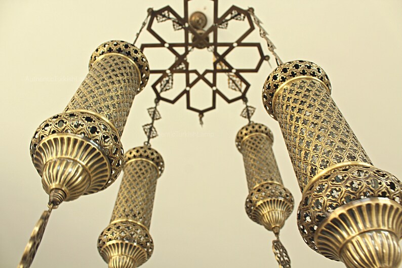 Puede incluir: L&aacute;mpara de ara&ntilde;a antigua y ornamentada con cinco l&aacute;mparas cil&iacute;ndricas doradas. Las l&aacute;mparas presentan intrincados dise&ntilde;os de metal calado y cuelgan de un accesorio de techo decorativo con un patr&oacute;n en forma de estrella. La est&eacute;tica general es vintage y elegante.