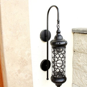 May include: Black metal outdoor wall sconce with a clear glass shade featuring a geometric pattern. The sconce has a curved arm and is mounted on a white wall.