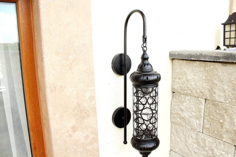 May include: Black metal outdoor wall sconce with a clear glass shade featuring a geometric pattern. The sconce has a curved arm and is mounted on a white wall.