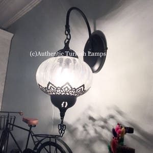 May include: A white glass Turkish wall lamp with a black metal frame and a chain hanging from the top. The lamp is lit and casting a warm glow. The lamp is mounted on a wall with a white background. (c)Authentic Turkish Lamps