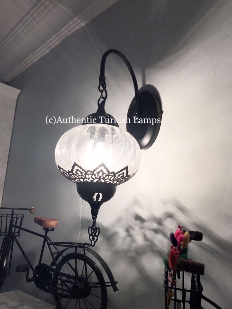 May include: A white glass Turkish wall lamp with a black metal frame and a chain hanging from the top. The lamp is lit and casting a warm glow. The lamp is mounted on a wall with a white background. (c)Authentic Turkish Lamps